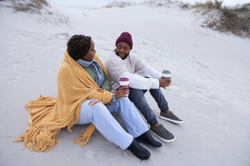Obraz premium African American couple sitting on sand dune wrapped in mustard blanket, maroon beanie