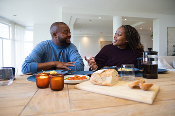 Obraz premium African American couple sitting at wooden dining table in home, chatting over bread and coffee