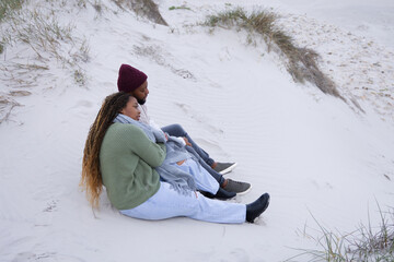Obraz premium African couple sitting on sand dune at beach wearing maroon beanie and green sweater, copy space