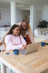 Obraz premium African American couple sitting at home at wooden dining table tapping silver laptop and blue mugs