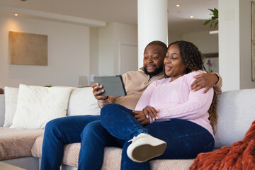 Obraz premium African American couple sitting on light-colored sectional sofa in modern living room, using tablet