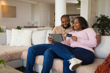 Fototapeta premium African couple sitting on sectional sofa in bright living room, holding tablet and credit card