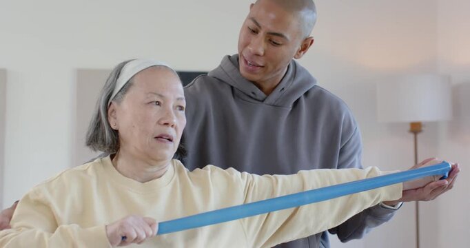 Senior Asian woman pulling blue band, being guided by African American aide at home for strength