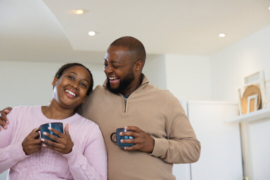 Couple holding dark mugs, leaning and laughing in kitchen wearing pink sweater and tan pullover