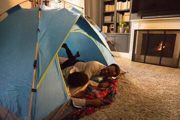 Obraz premium African American father and child lying in blue tent on gray rug in living room, talking