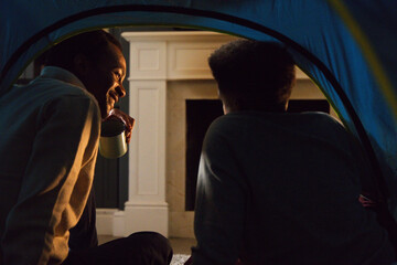 Obraz premium Two African American males, one teenage, sitting in blue tent by white mantel fireplace holding mug