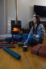 Obraz premium Lady sitting at home wearing sweater and festive headband wrapping gifts, mug near stove