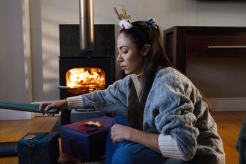 Obraz premium Woman sitting on wood floor near stove, fire glowing, in sweater reaching wrapping roll with gifts