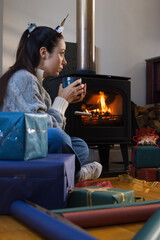 Obraz premium Woman wearing knit sweater holding mug sitting by wood stove with wrapped gifts and paper