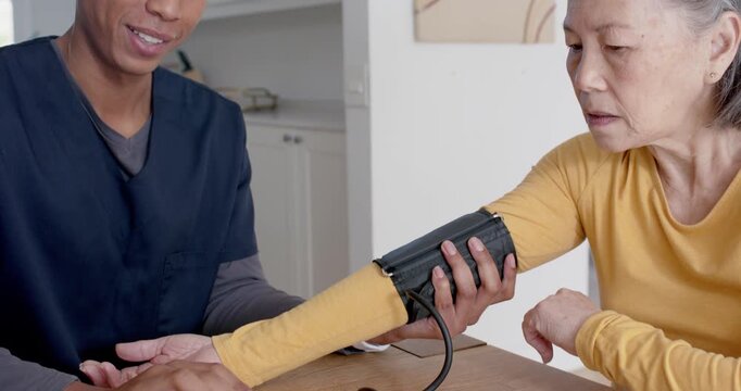 Senior Asian woman after placing cuff, African American nurse checking BP and reassuring at kitchen