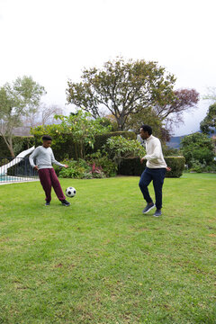 African American teenager and man kicking soccer ball on lawn near fenced pool with white slide