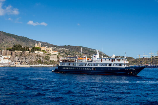 Lipari, ME - IT - Sep 06, 2026 : Passenger ship Artemis crosses blue harbor water off Lipari, with the historic hilltop church complex and Star Clippers sailing ship visible behind. under clear skies.