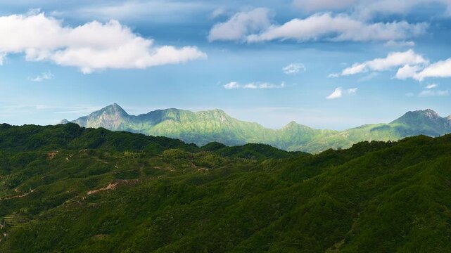 Lush green mountain peaks rise under a blue sky with fluffy white clouds, illuminated by bright daylight across the scenic landscape.