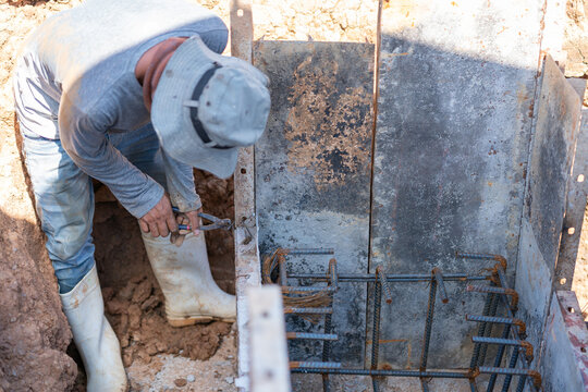 High-angle view of a builder tying steel reinforcement bars with binding wire in a pit for concrete footing installation.