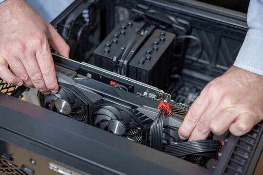 A technician installs a powerful triple-fan gaming graphics card on a gaming PC motherboard. A dual-tower CPU cooler and black power cables are visible