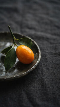 A fresh whole kumquat with leafy twig on dark ceramic dish over linen