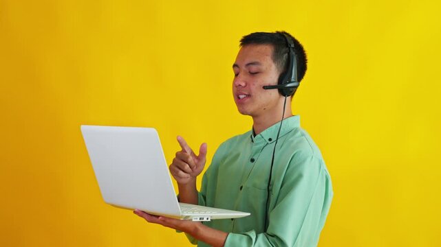 Excited Asian headset operator holding laptop computer, waving hand, and typing on bright yellow studio backdrop