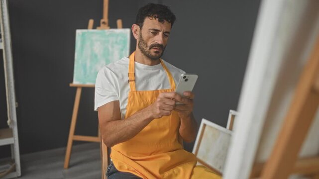 Man painter in yellow apron seated tapping smartphone with fingers while looking aside at canvas and easel in studio; quiet contemplation.