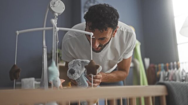 Man leans over wooden crib adjusting hanging mobile and plush bunny with hand in building nursery while peering into bed; bonding tenderness.