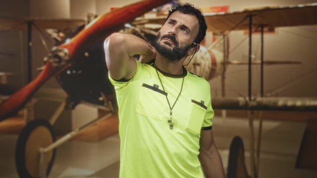 Man referee middle aged bearded hispanic with whistle and headset, hand behind neck stretch in a building with vintage biplane exhibit; tired reflection.