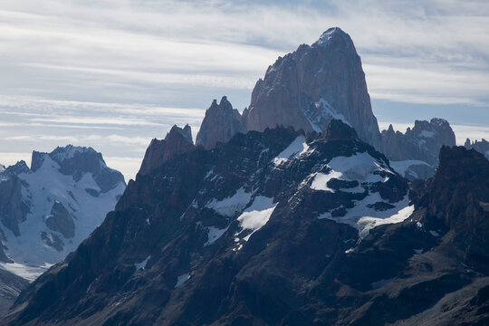Majestic granite mass of Mount Fitz Roy rising above dark jagged ridges and snowfields under a cloudy sky in Los Glaciares National Park, Patagonia.
