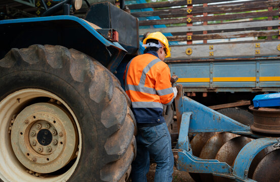 Agricultural mechanic inspects a tractor and farm equipment while wearing safety gear, showing machinery maintenance, preventive service, technical work, and reliable agricultural operations.
