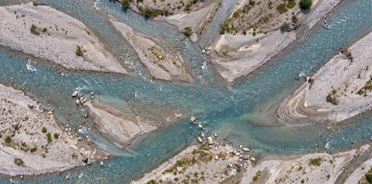 Aerial view of a braided river system with turquoise water flowing through gray gravel bars, sediment deposits, and scattered rocks in a rugged mountain landscape