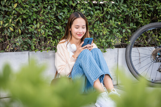 In a serene park on a lovely spring day a cute woman sits with her bicycle happily using her mobile phone to listen to music with earphones. Her joyful typing and bright smile showcase joy of season.