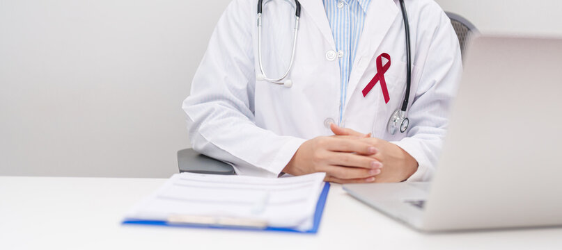 doctor using laptop and report notes on notebook. burgundy ribbon concept of March Multiple Myeloma Cancer Awareness month, September Sickle Cell Anemia, Headache and Migraine week, April Meningitis