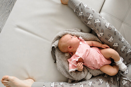 Baby 5-6 months old in pink clothes lies on sofa between mother's legs while she sits nearby, photographed from above at home