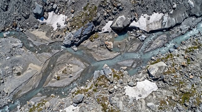 Aerial view of a glacial meltwater stream winding through a rocky mountain landscape with patches of snow and grey stone debris, captured from a perspective
