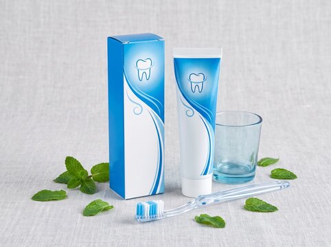 Blue toothpaste tube and box with a tooth logo on a linen surface. Includes a toothbrush, empty glass, and fresh mint leaves for oral hygiene.