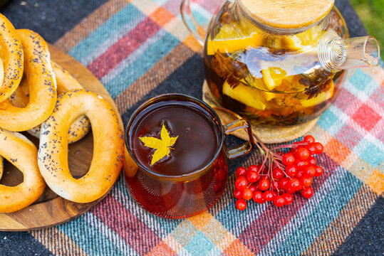 Glass cup filled with hot autumn tea featuring floating yellow maple leaf as central focus during outdoor picnic. Transparent teapot red viburnum berries and traditional bagels rest nearby on rustic