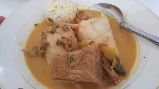 Katupek Pitalah is a traditional Minangkabau dish from Padang Panjang, West Sumatra, featuring rice cake, aged jackfruit curry, boiled egg, noodles and aromatic herbs on a plate with spoon. Closeup