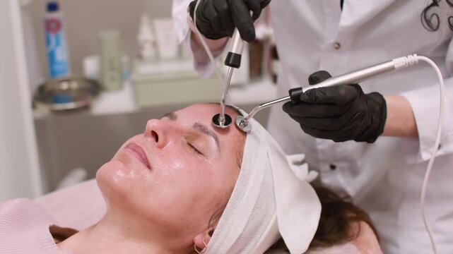Face lifting. Aesthetic therapy. Female in black gloves using professional device for modern microcurrent beauty procedure in salon.