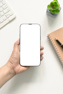 Top view flat lay hand holding modern smartphone with blank white screen template, keyboard, notebook, pen, and plant on white desk. Concept for marketing, website, branding, social.