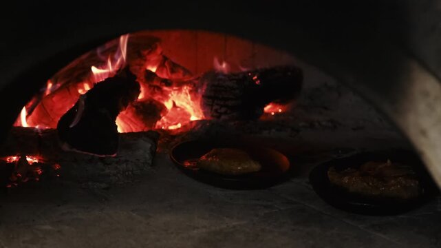 Roasting food. Vintage stone oven. Black pan with meat meal baking in traditional arch brick fireplace with burning flame on dark logs.