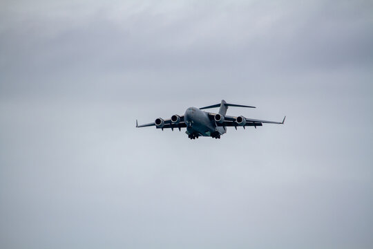 C-17 Globemaster III military transport aircraft photographed in flight against an overcast sky, viewed from below with four jet engines, high-mounted wings and landing gear extended.