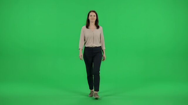 Full body shot of a confident young professional woman in business casual attire walking slowly forward towards the camera against a solid green screen.