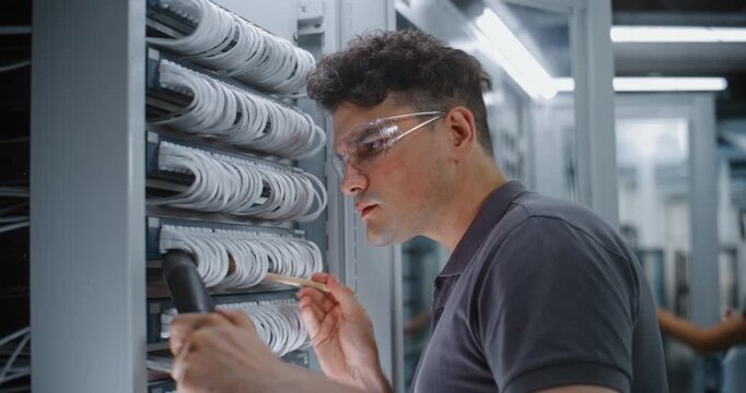 Male Network Technician Wearing Protective Glasses Carefully Cleans Delicate Ethernet Connections on Server Rack Using Brush and Vacuum Hose. Detailed Hardware Servicing Optimize Signal Integrity.