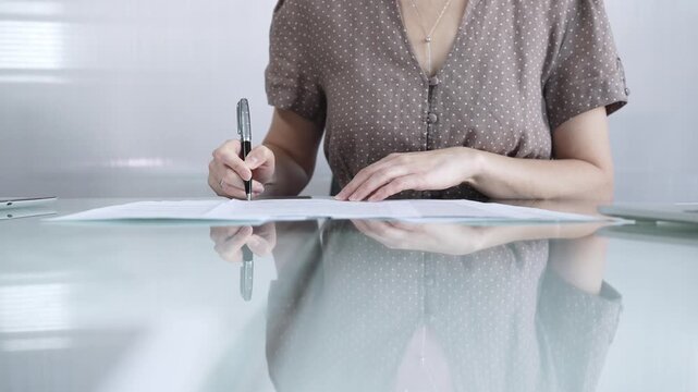 Businesswoman in brawn dotted blouse is signing a contract at a sleek glass desk in a modern office, showcasing professionalism and focus in her work environment, closeup. Business people concept