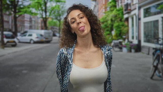 Young hispanic woman in white tank top and patterned open shirt showing bare chest, smiling and sticking tongue out while standing on a city street; playful confidence.