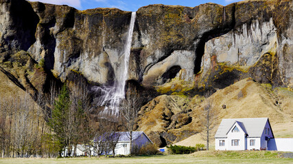 A slender waterfall cascades down a sheer, cavernous cliff face directly behind rural cottages and farmhouses in southern Iceland © Gregory