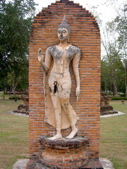 タイのスコータイ歴史公園にあるレンガ壁の前に立つ優美なスコータイ様式の遊行仏立像 © Zumi