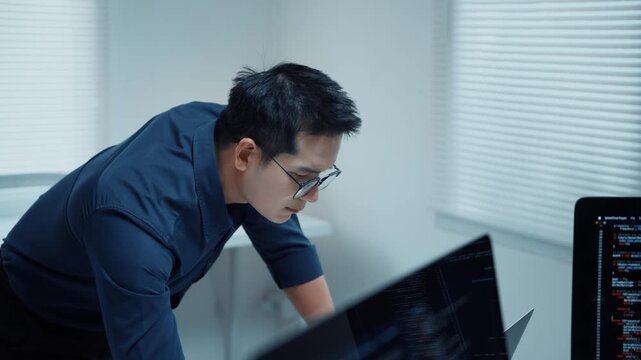 Focused Asian programmer thinking while developing software and coding on a laptop in a modern office with multiple displays