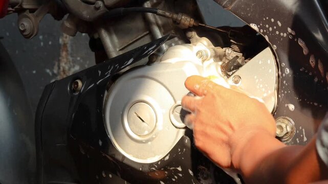 Close-up of a person's hand cleaning a motorcycle engine and body with a soapy sponge during vehicle maintenance and washing at home.
