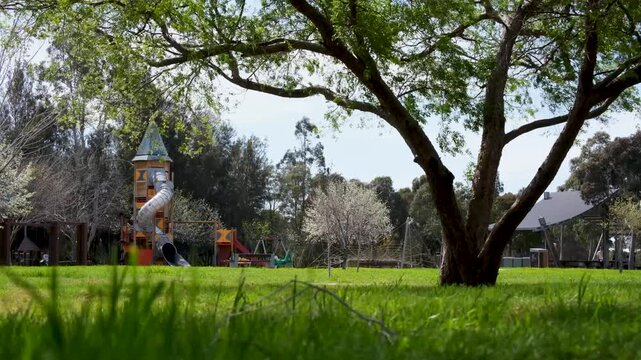 Distant view of children playing at a suburban park playground with a rocket slide tower and mature shade trees in Melbourne's western suburbs, Sunshine west, Victoria, Australia