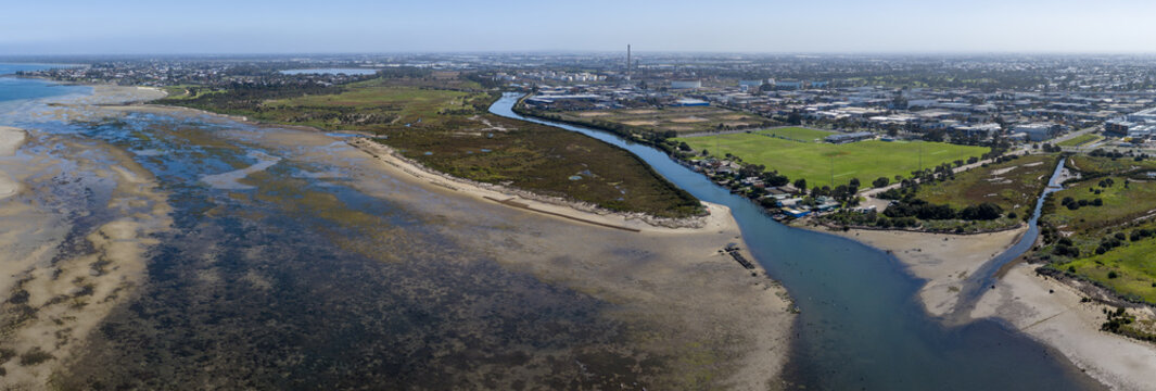 Aerial panorama of Kororoit Creek mouth entering Port Phillip Bay with tidal mudflats, saltmarshes, and industrial background in Altona and Williamstown, Melbourne, Victoria, Australia