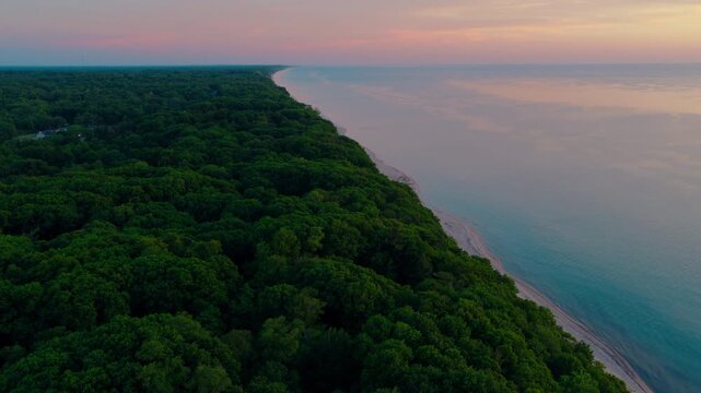Aerial over Michigan wilderness at dusk, where hardwood forest covers a bluff above an empty stretch of sand beach and calm Lake Michigan water.