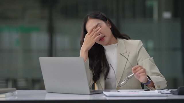 Stressed asian businesswoman experiencing headache and eye strain from overworking on her laptop in a modern corporate office